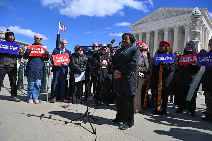 A group of people holding signs and speaking in front of a large, white building with pillars.