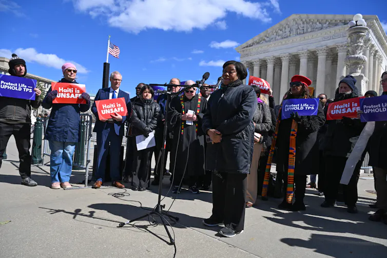 A group of people holding signs and speaking in front of a large, white building with pillars.