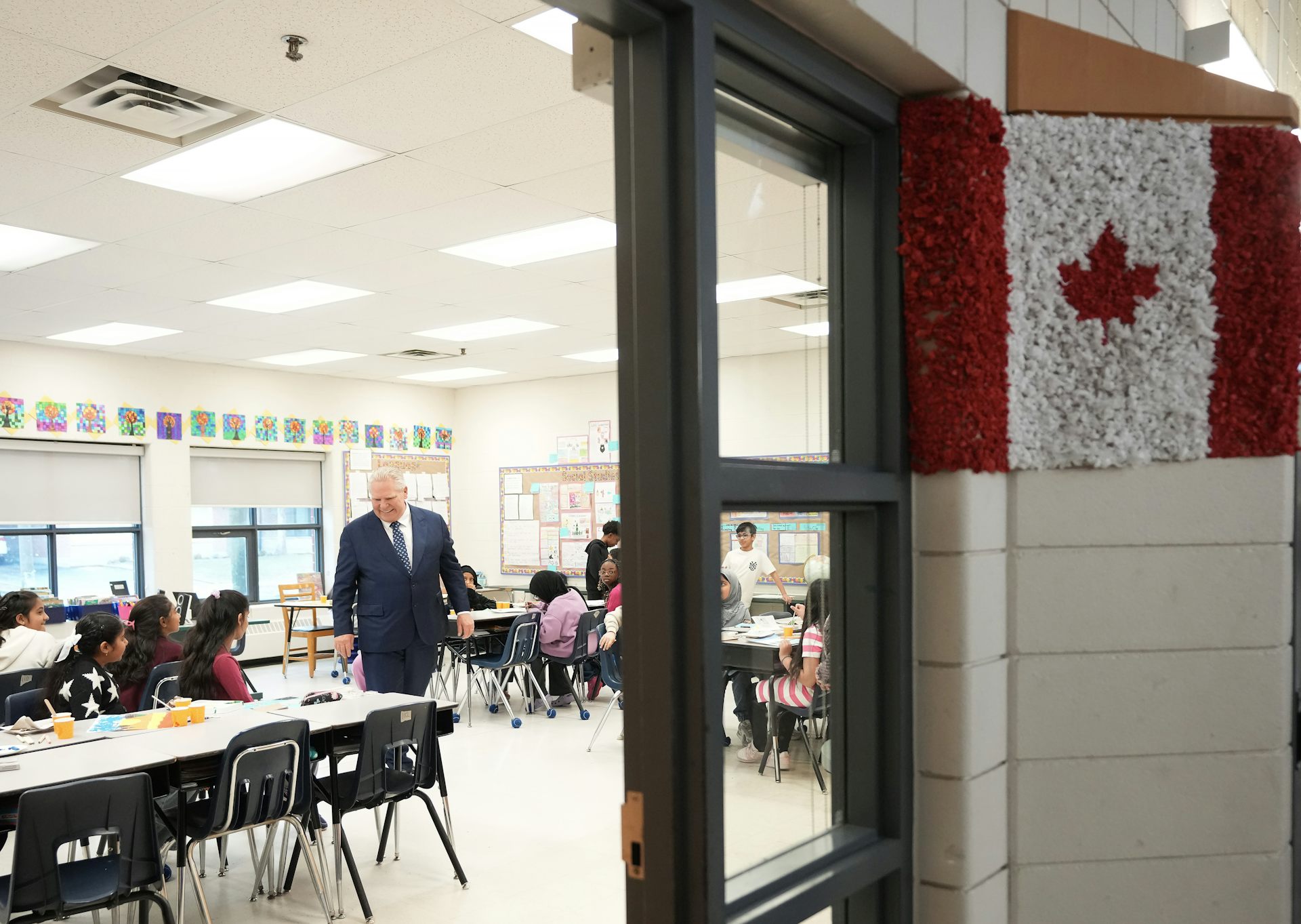 A man in a suit stands in a classroom near a Canadian flag made of tissue paper.
