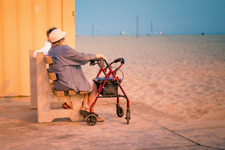 An older woman with a walking frame sits on a public bench on a beach.