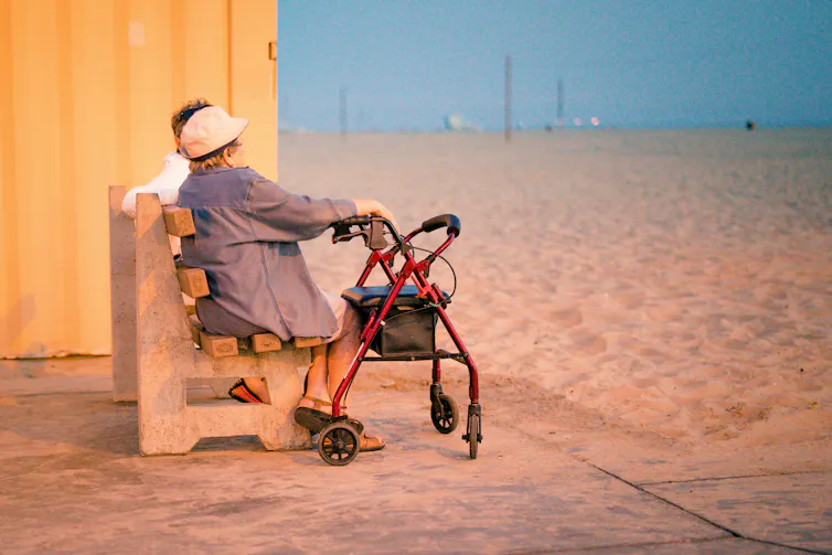An older woman with a walking frame sits on a public bench on a beach.