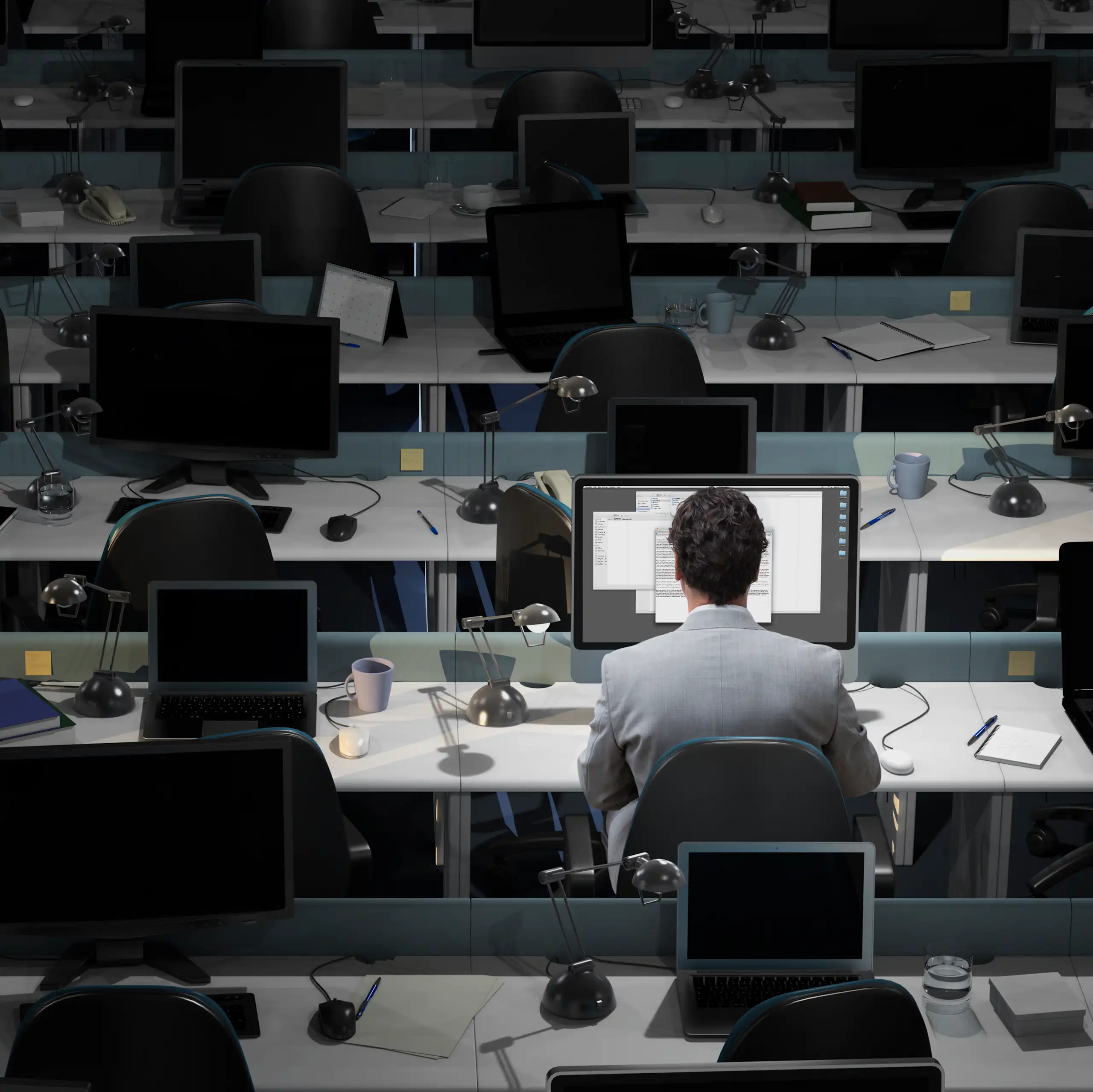 A man sits at a computer, alone in an open plan office filled with desks.