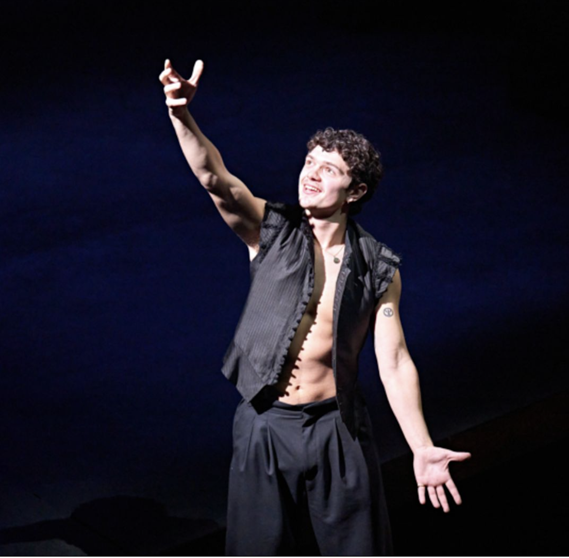 A young man on a black stage background dressed in black arms aloft looking happy.