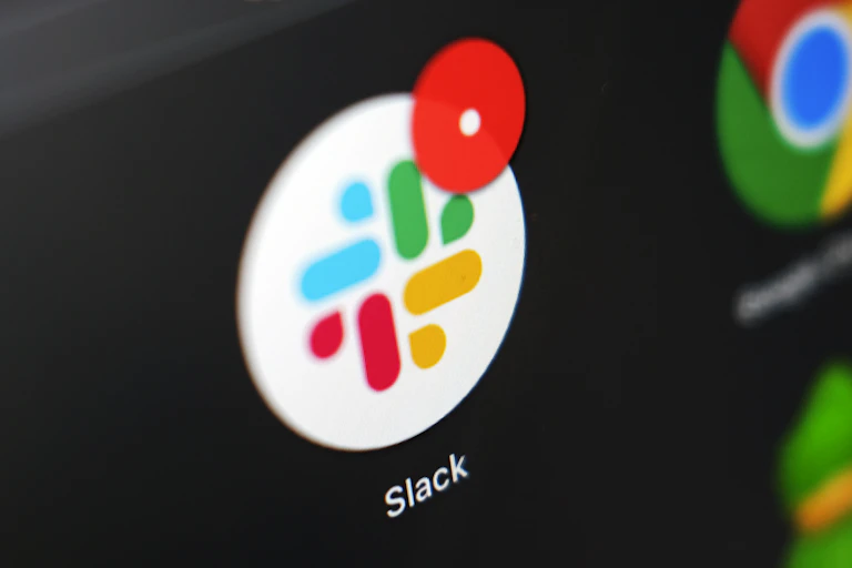 A Slack icon, with its trademark blue, green, yellow and red pattern, is displayed against a dark screen with a message notification on its upper right.
