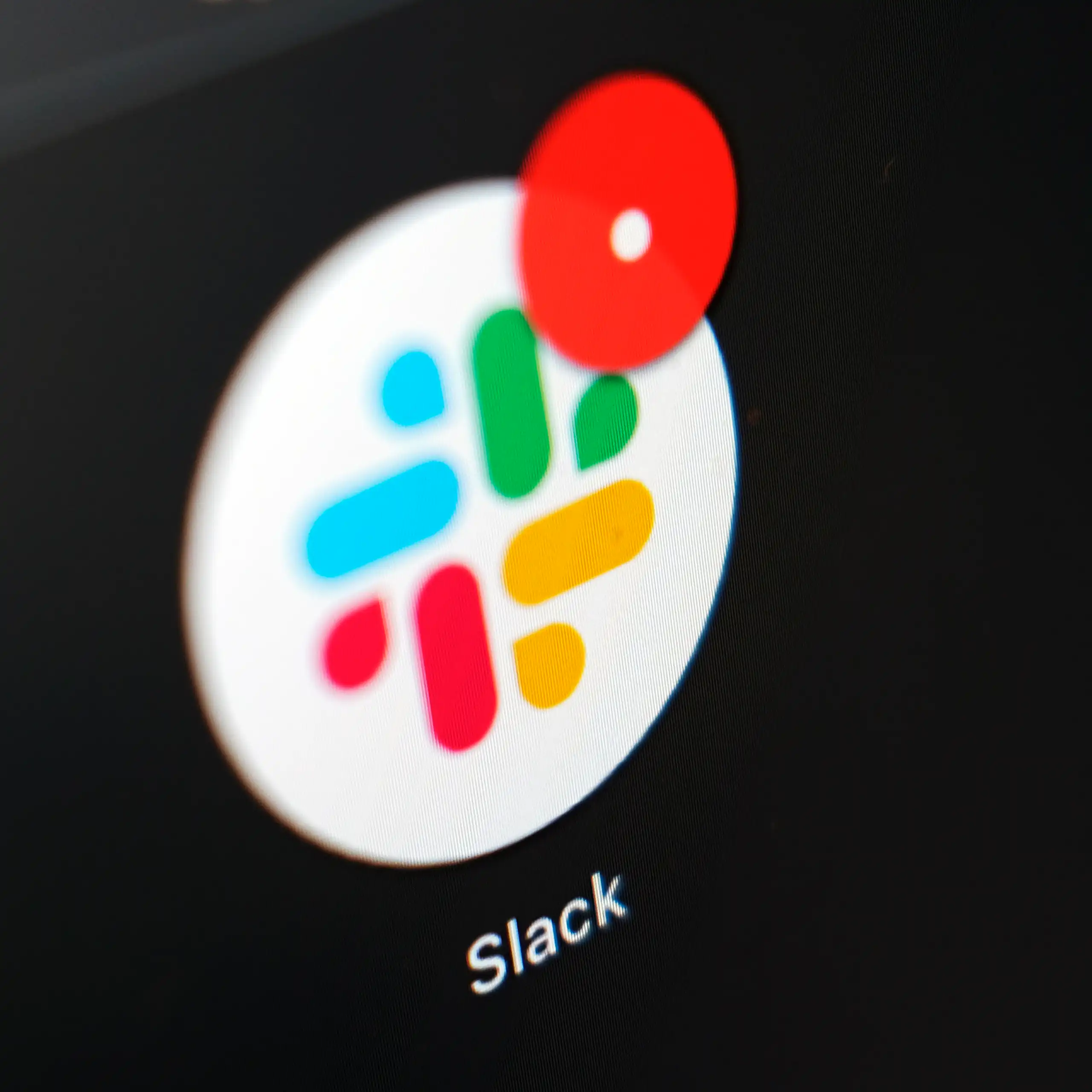 A Slack icon, with its trademark blue, green, yellow and red pattern, is displayed against a dark screen with a message notification on its upper right.