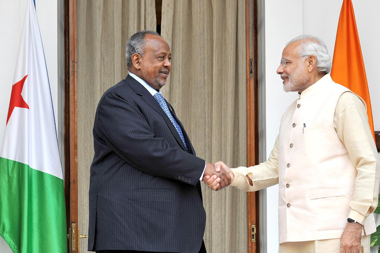 Two men standing and shaking hands in front of flags positioned behind each of them