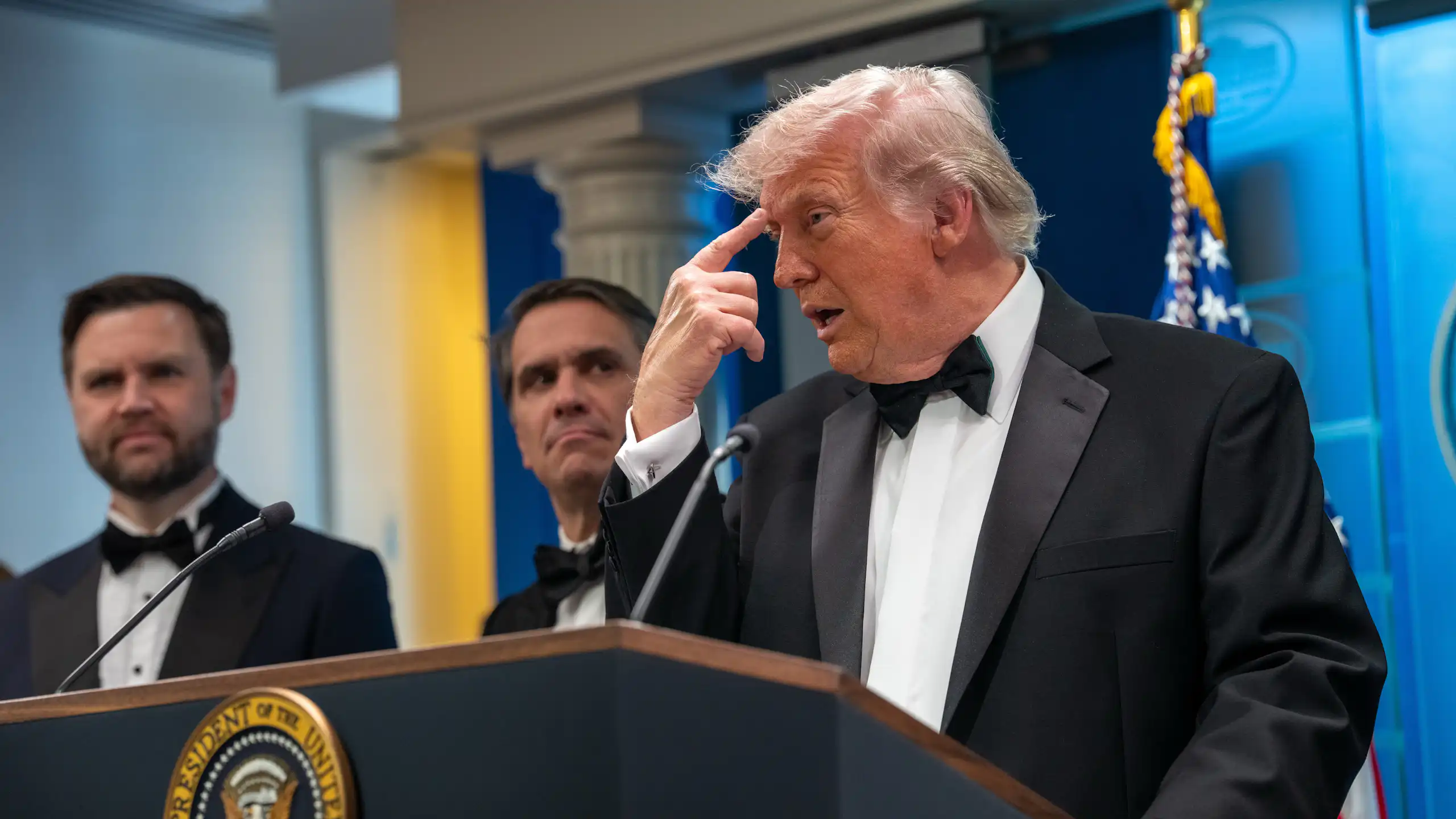 A man in a suit standing in front of a podium points to his head.