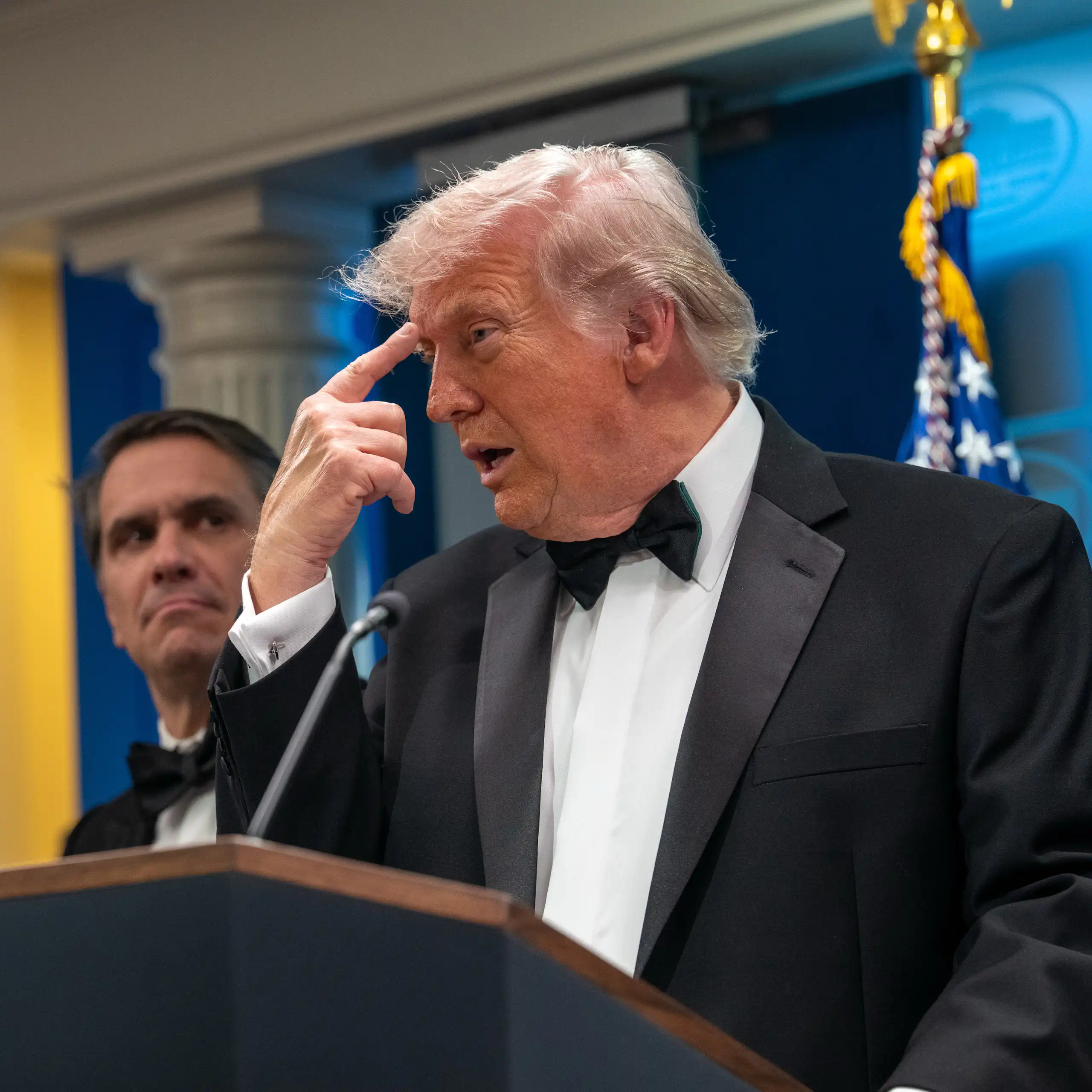A man in a suit standing in front of a podium points to his head.
