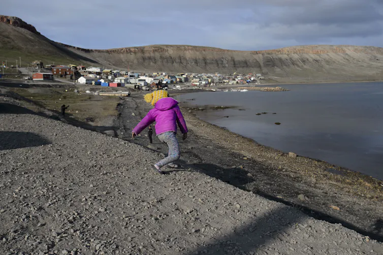 A child in a pink jacket throws a stone into a body of water. Grey hills and a coastal village are in the background.