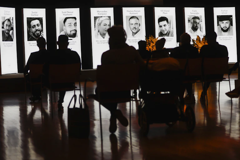 A group of people gathering to remember nine people that were killed in a shooting. They are sitting in front of large scale headshots of the victims..