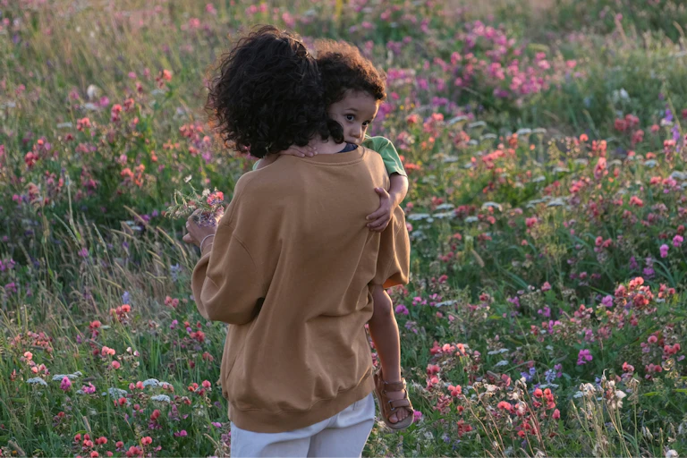 A woman walks through a field with wildflowers holding a toddler.