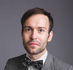man with brown hair, dressed in suit and bow tie, grey background