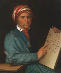 A painted portrait of a man in a blue coat and red turban smoking a pipe, pointing to a chart of letter-like symbols.