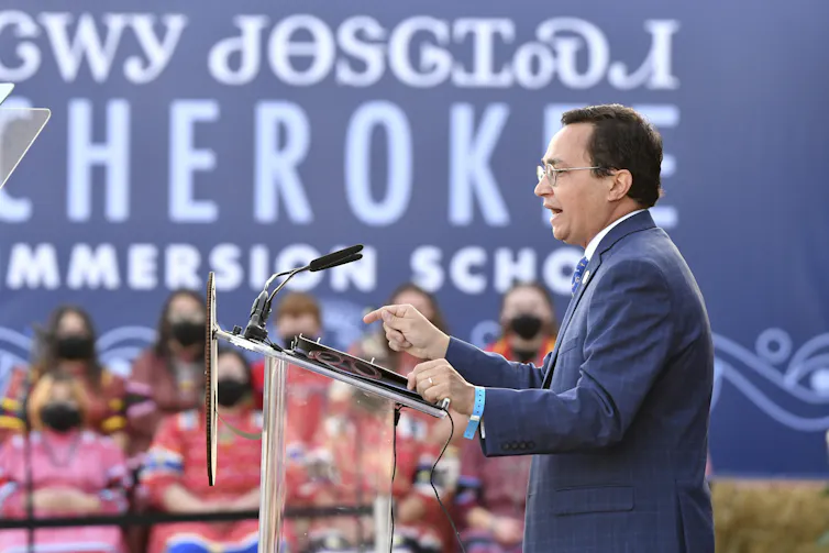 La Biblia Cherokee, uno de los primeros libros en el idioma, es una ventana entre visiones del mundo 1 Un hombre con traje azul habla en un atril mientras mujeres vestidas de rosa brillante y rojo se sientan detrás de él.