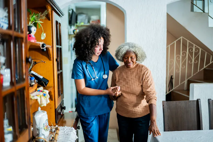 A home health aide helps a patient walk around a private home.