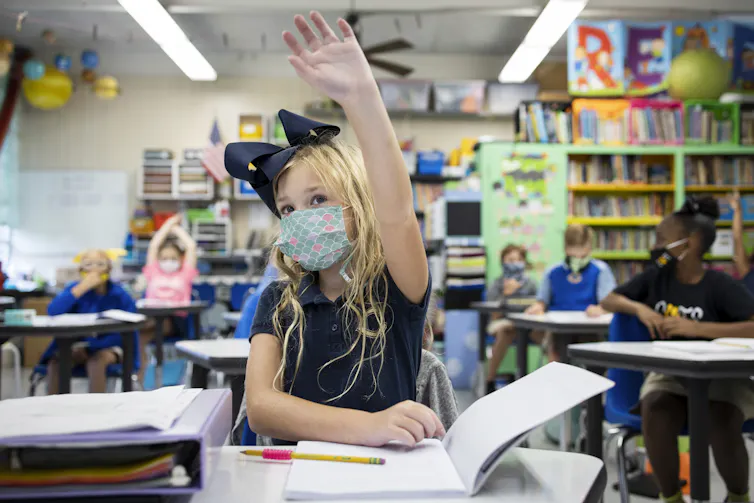 A girl with blonde hair and a large bow wears a face mask and raises her hand, while she sits at her desk in a classroom with other students and books.