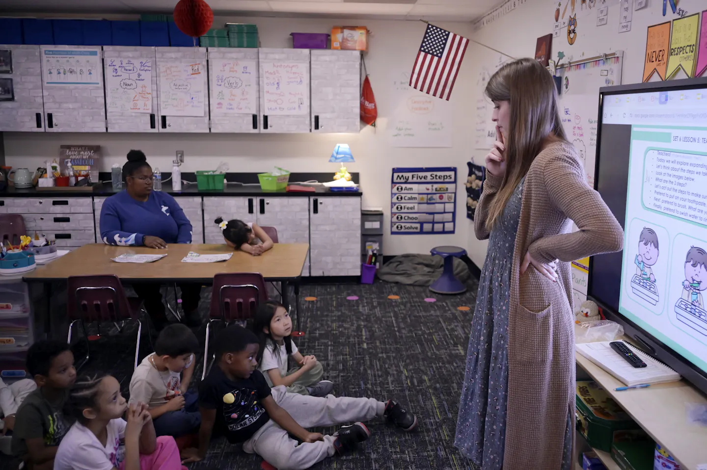A woman stands near a projector screen in front of a group of children seated on the floor in a classroom.