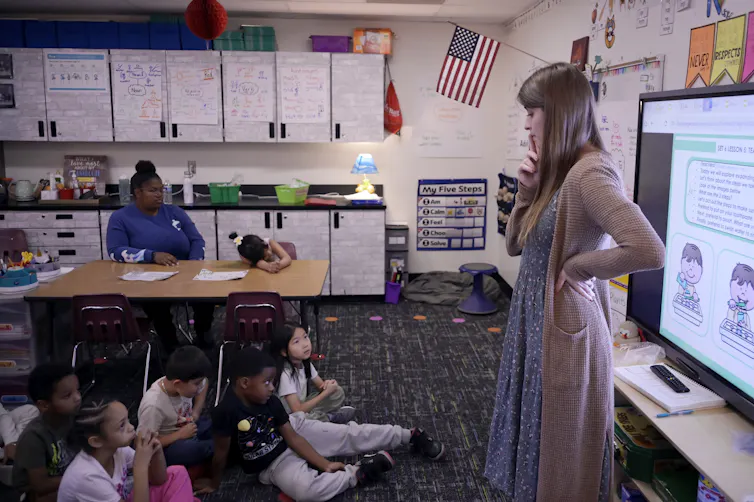Los avances en lectura en Alabama, Mississippi y Luisiana a menudo se promocionan, pero no muestran el panorama completo de la alfabetización. 1 Una mujer está parada cerca de una pantalla de proyección frente a un grupo de niños sentados en el suelo del aula.
