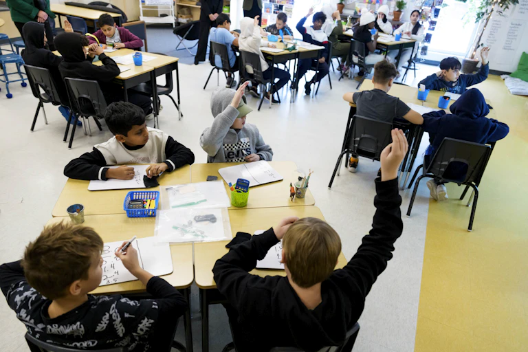 Students seen at tables and desks and one has a hand raised.