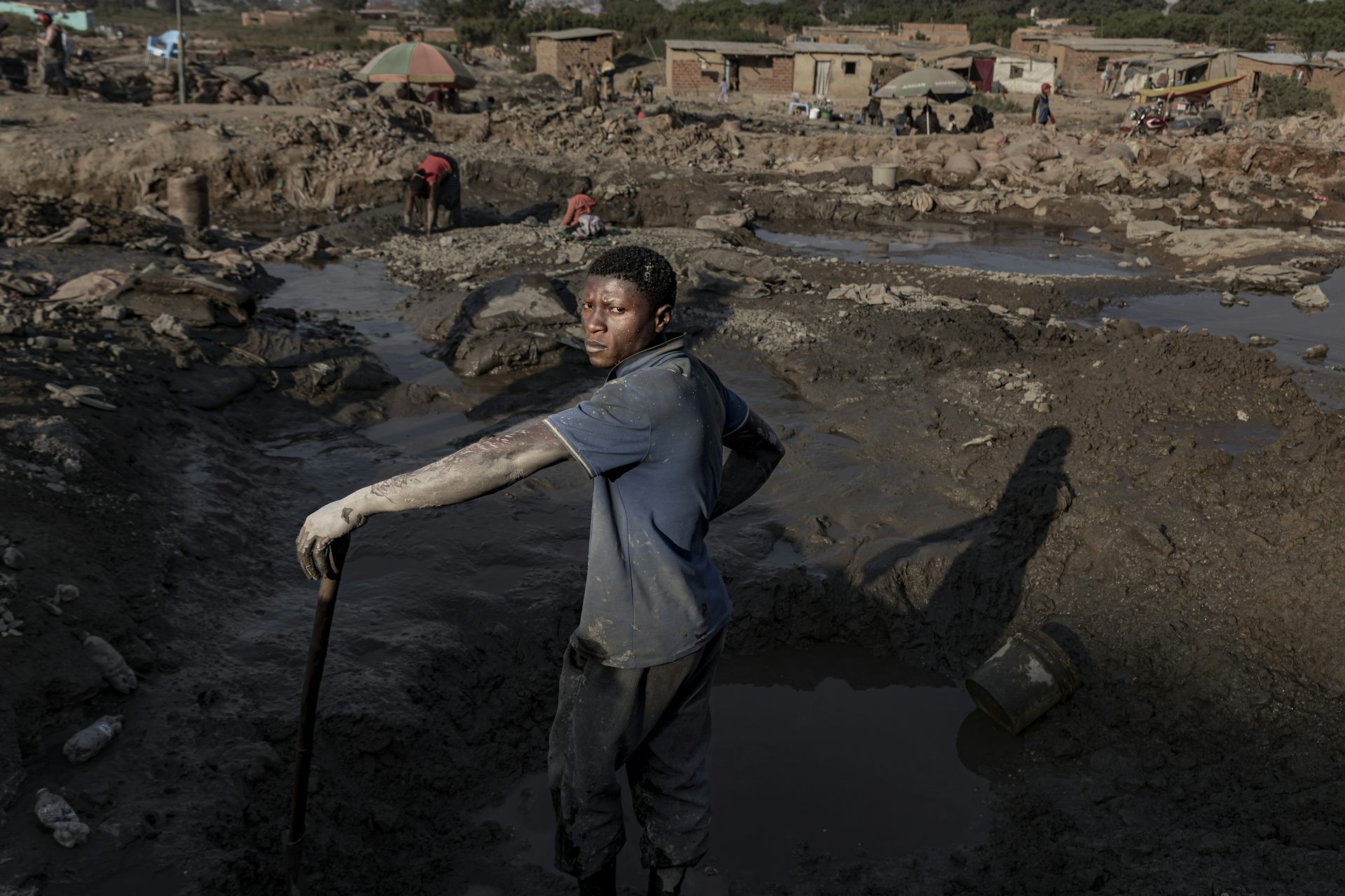 Un hombre con barro seco en sus manos desnudas se encuentra cerca de una mina llena de agua donde un niño y una mujer buscan minerales.
