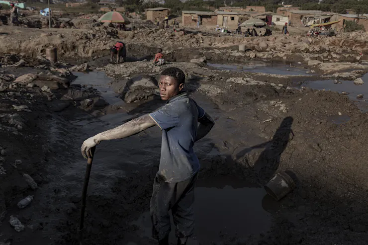 A man with dried mud on his bare arms stand near a water-filled mine where a child and woman are searching for minerals.