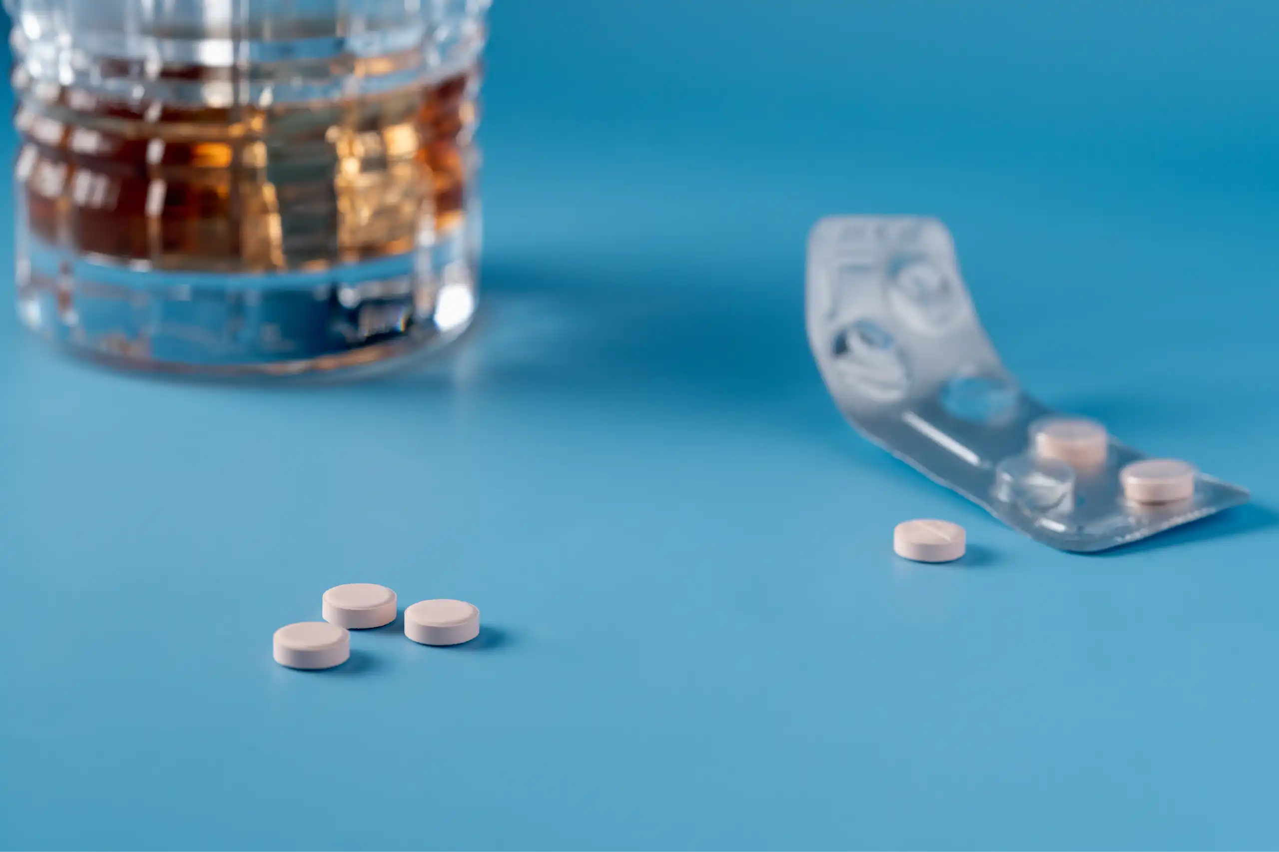 Photo d’un verre d’alcool posé à côté de médicaments, sur une table de couleur bleue.