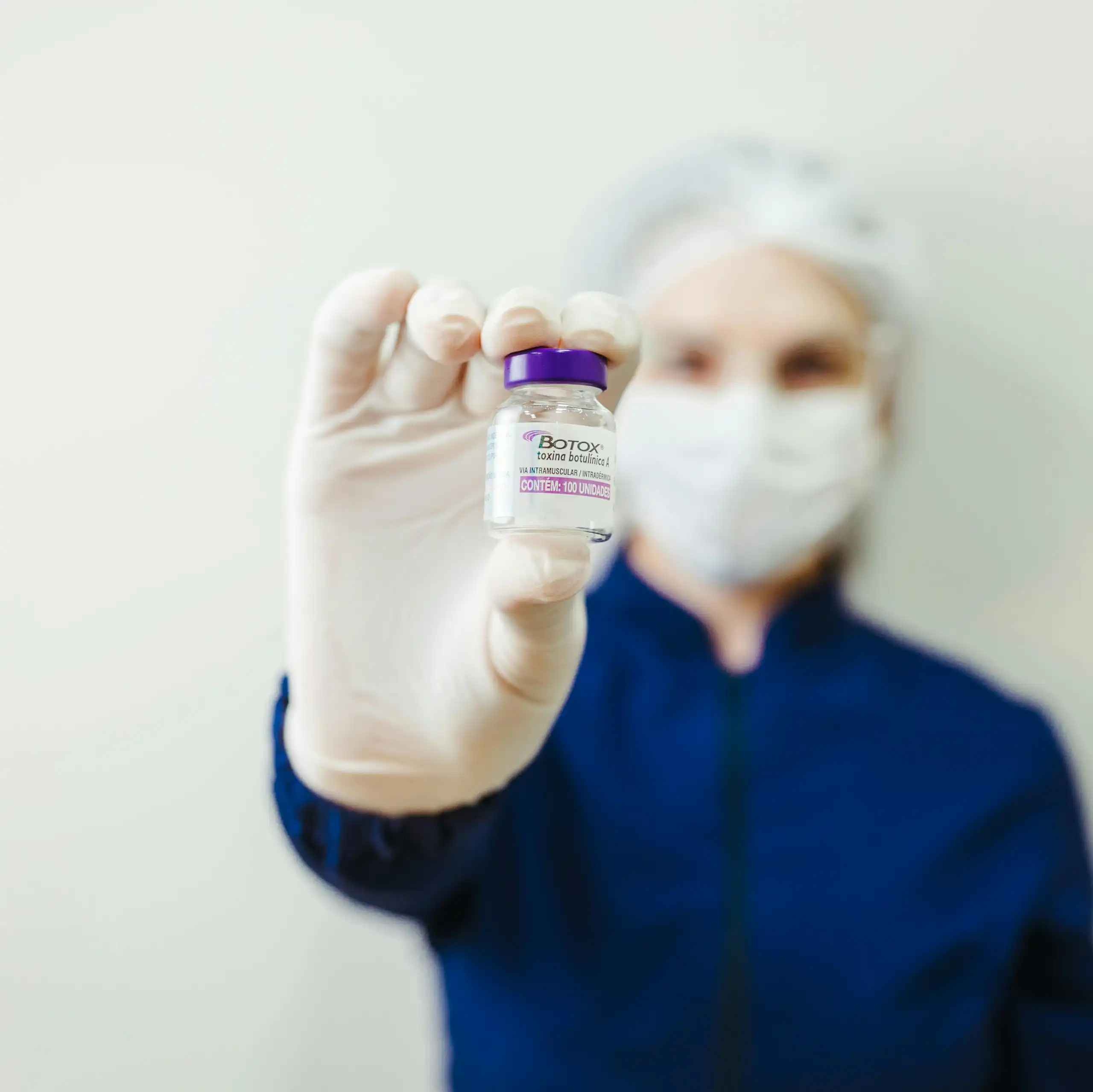 A woman in a mask and surgical gloves holding out what looks like a vial of Botox