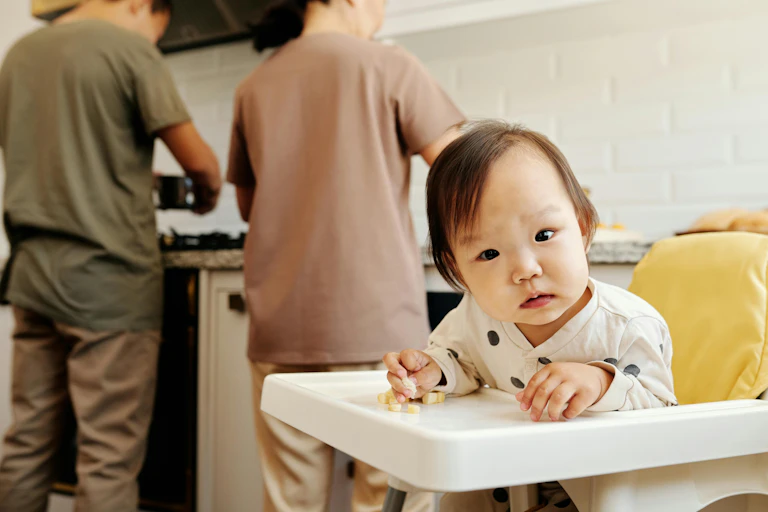 A curious baby sits in a high chair. Two adults with their backs turned work in a kitchen.