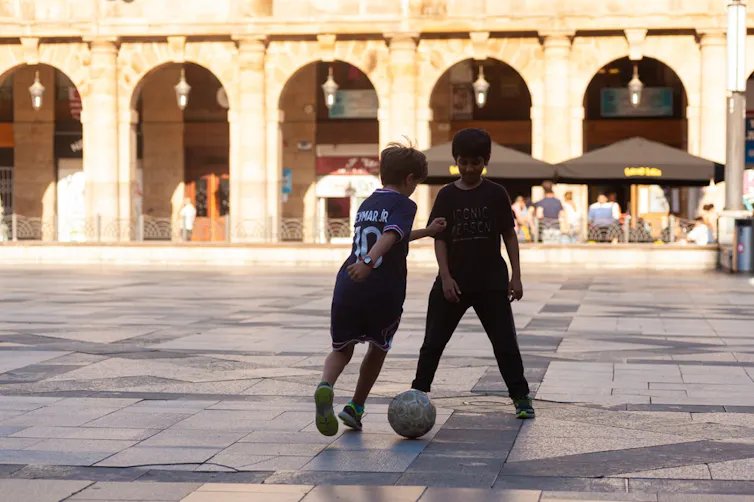 Dos niños juegan al fútbol en una plaza, a contraluz.