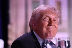 An older man with a fluffy white-blond hairstyle grins smugly.