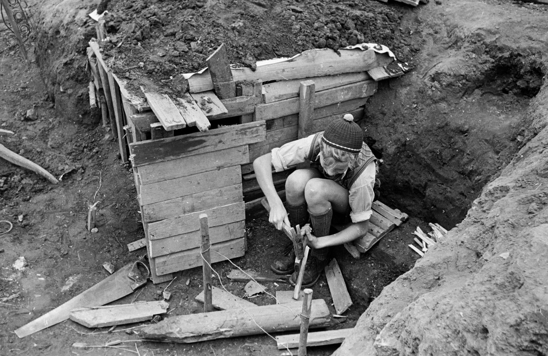 Black and white photo of boy kneeling in a ditch and using a hammer.