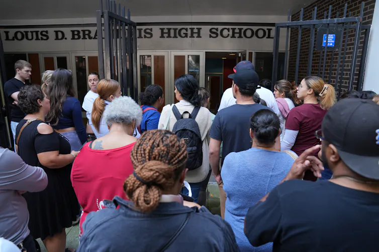 A group of adults stand together outside the doors of a building that says Brandeis High School.