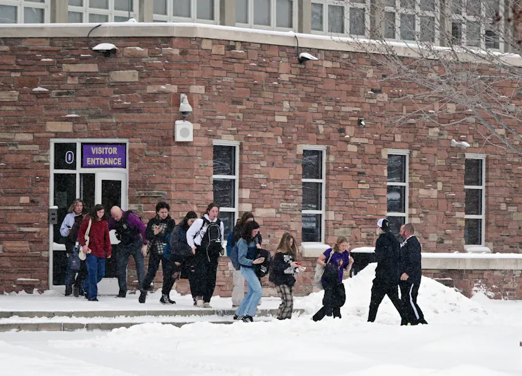 A group of people are seen exiting a brick building and walking along a snowy lawn.