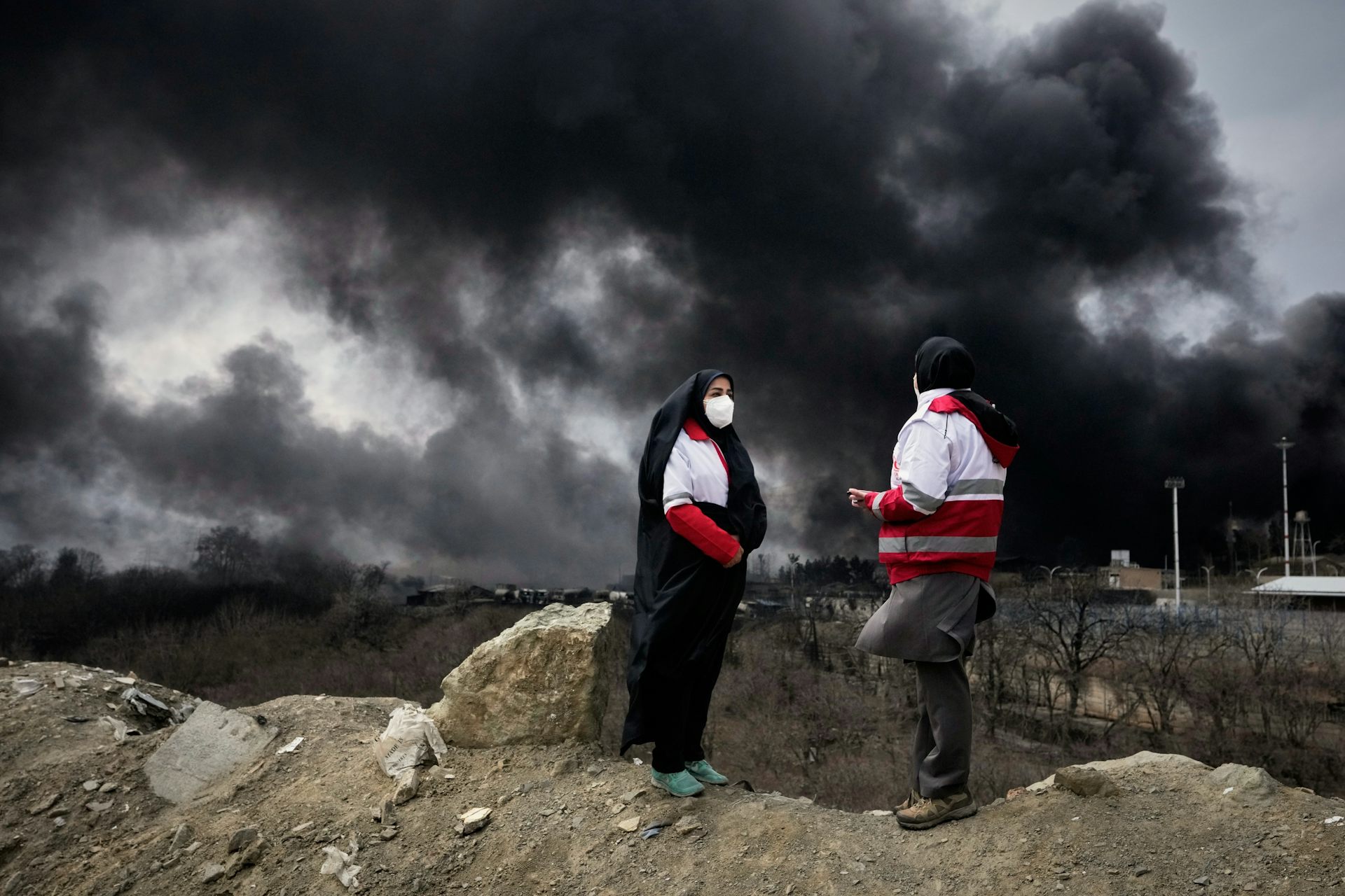 dos mujeres con hijabs y chaquetas rojas y blancas con máscaras faciales, grandes columnas de humo negro elevándose al fondo