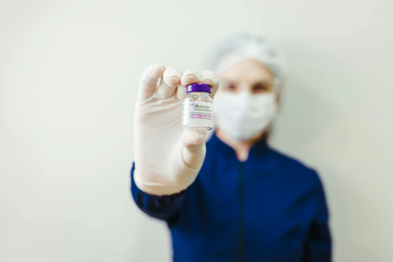 Uma mulher usando máscara e luvas cirúrgicas, segurando o que parece ser um frasco de Botox