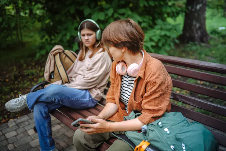 A teenage girl wearing headphones sits on a bench, looking toward another girl with her headphones around her neck a few inches away.