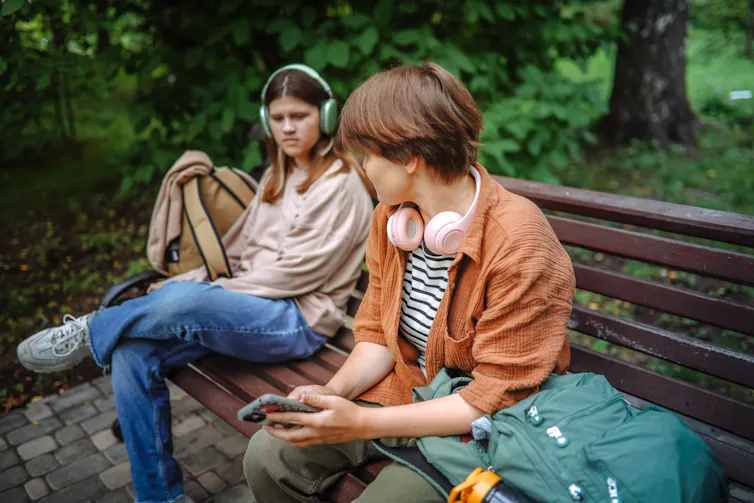A teenage girl wearing headphones sits on a bench, looking toward another girl with her headphones around her neck a few inches away.