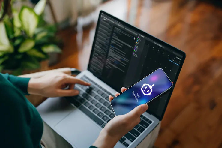 Someone whose face is out of the frame types with one hand on a laptop while holding up a laptop with a chatbot screen.