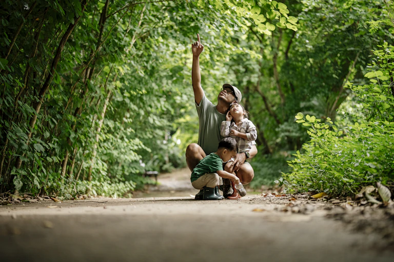 Father kneels with two young kids on a path in the forest, pointing to something up above.