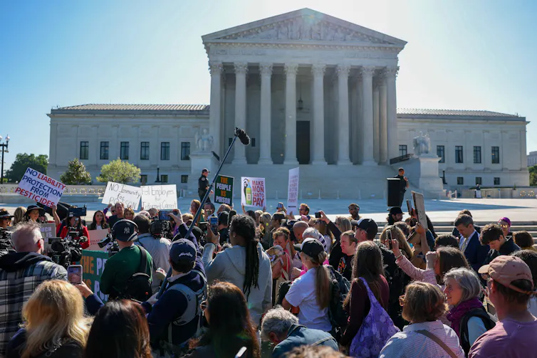 A group of people gather in front of a large white building with pillars in the front.