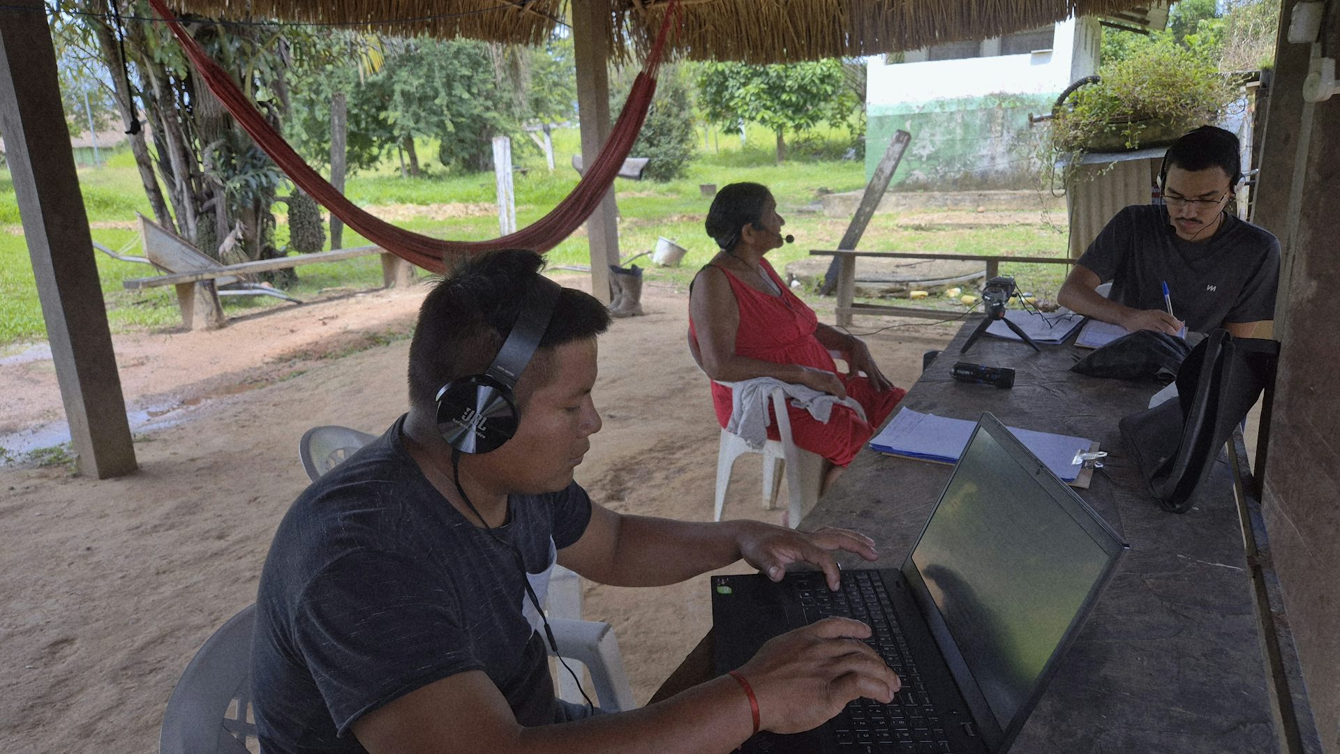 Foto mostra aldeia indígena, com rede ao fundo. Numa mesa, dois jovens com computadores e fones de ouvido conversam com uma mulher mais velha, que também usa fone de ouvido