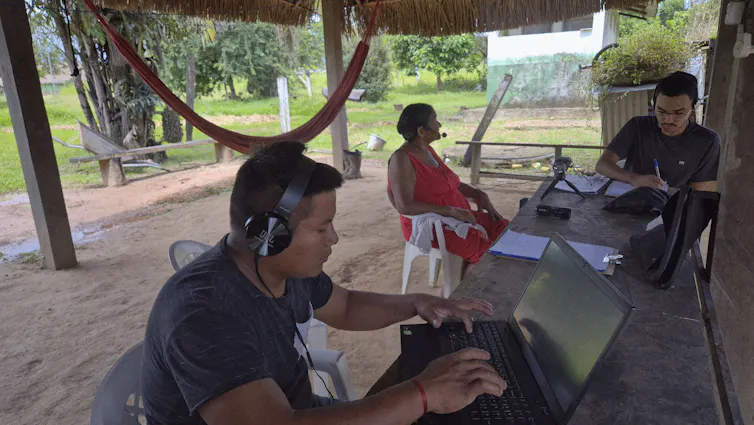 Foto mostra aldeia indígena, com rede ao fundo. Numa mesa, dois jovens com computadores e fones de ouvido conversam com uma mulher mais velha, que também usa fone de ouvido