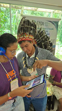Foto mostra um indígena, vestindo calça jeans e camiseta, com cocar e colar, mostrando um ipad para uma mulher, que segura o ipad e usa fones de ouvido