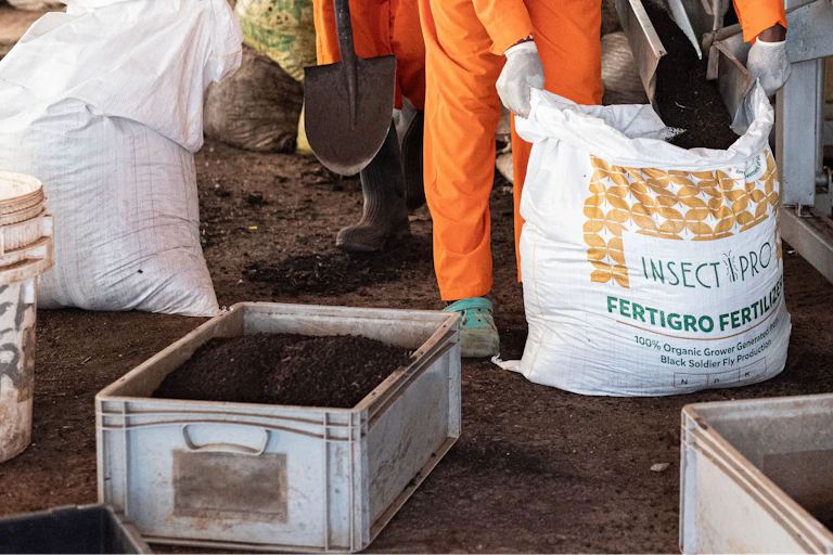A man stands with a sack bearing the word 'Fertiliser' next to a conveyor belt of organic matter.
