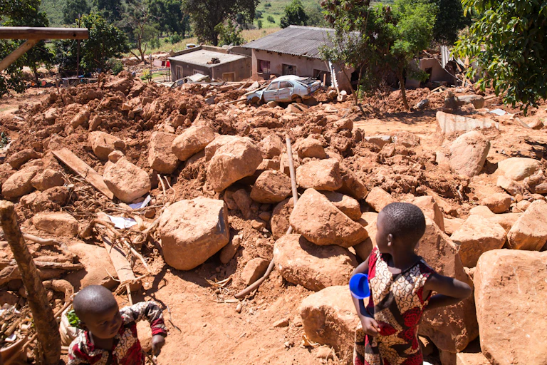 A small child looks out across a landscape that is covered by giant boulders and mud washed over houses and cars