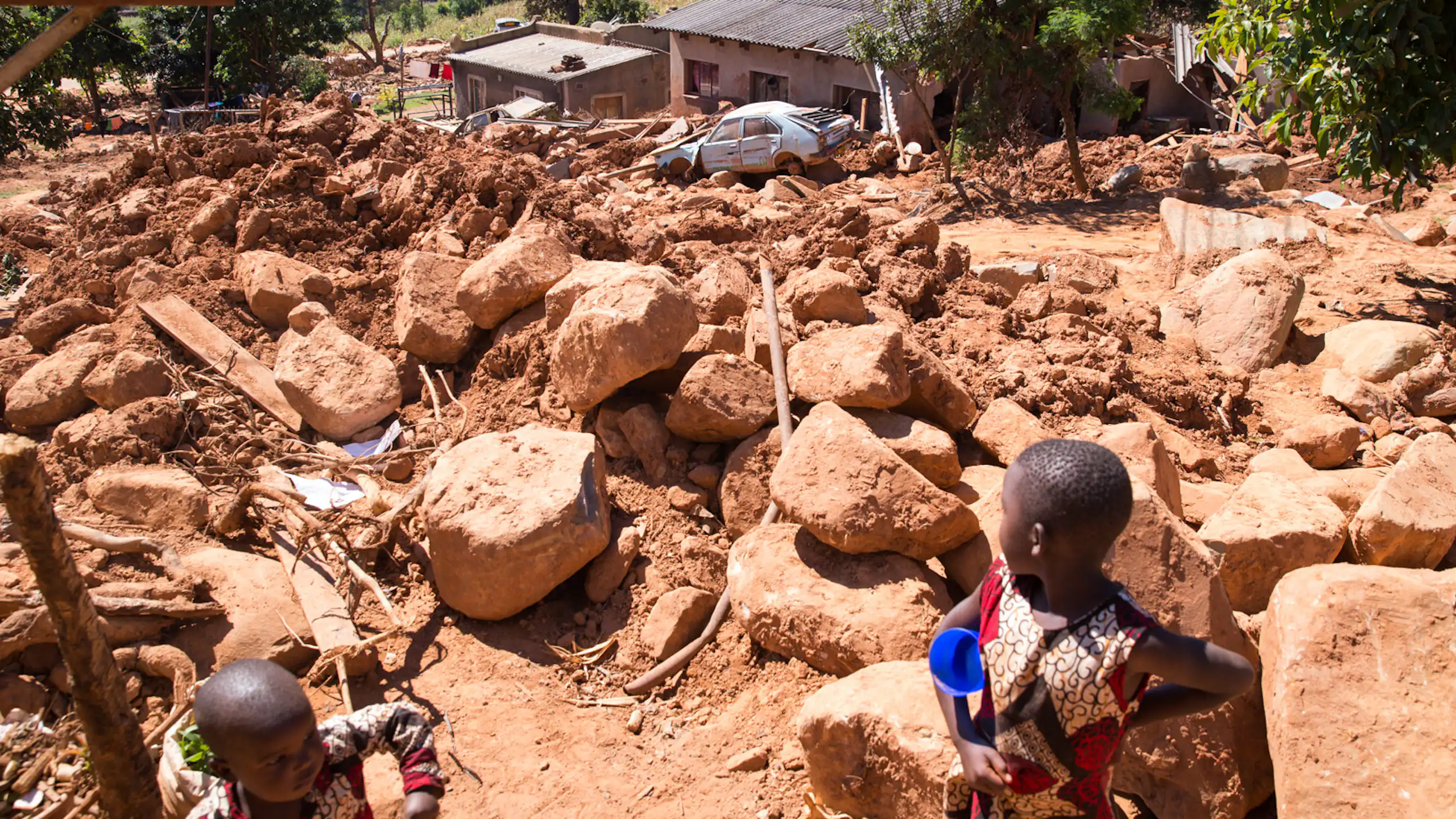 A small child looks out across a landscape that is covered by giant boulders and mud washed over houses and cars