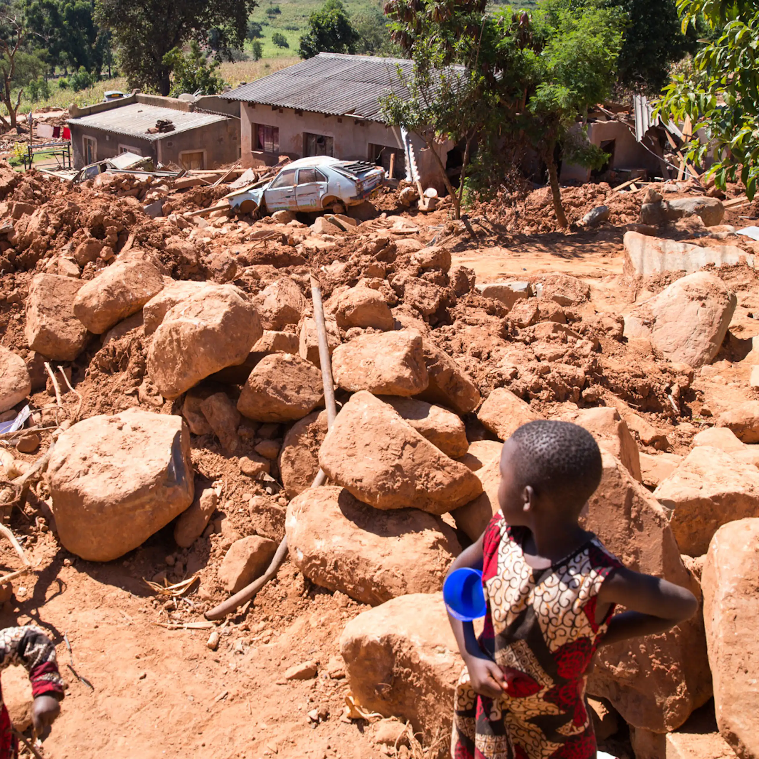 A small child looks out across a landscape that is covered by giant boulders and mud washed over houses and cars