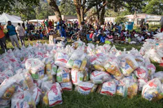 Dozens of people wait under trees for some basic food supplies which are piled up on the ground in front of them