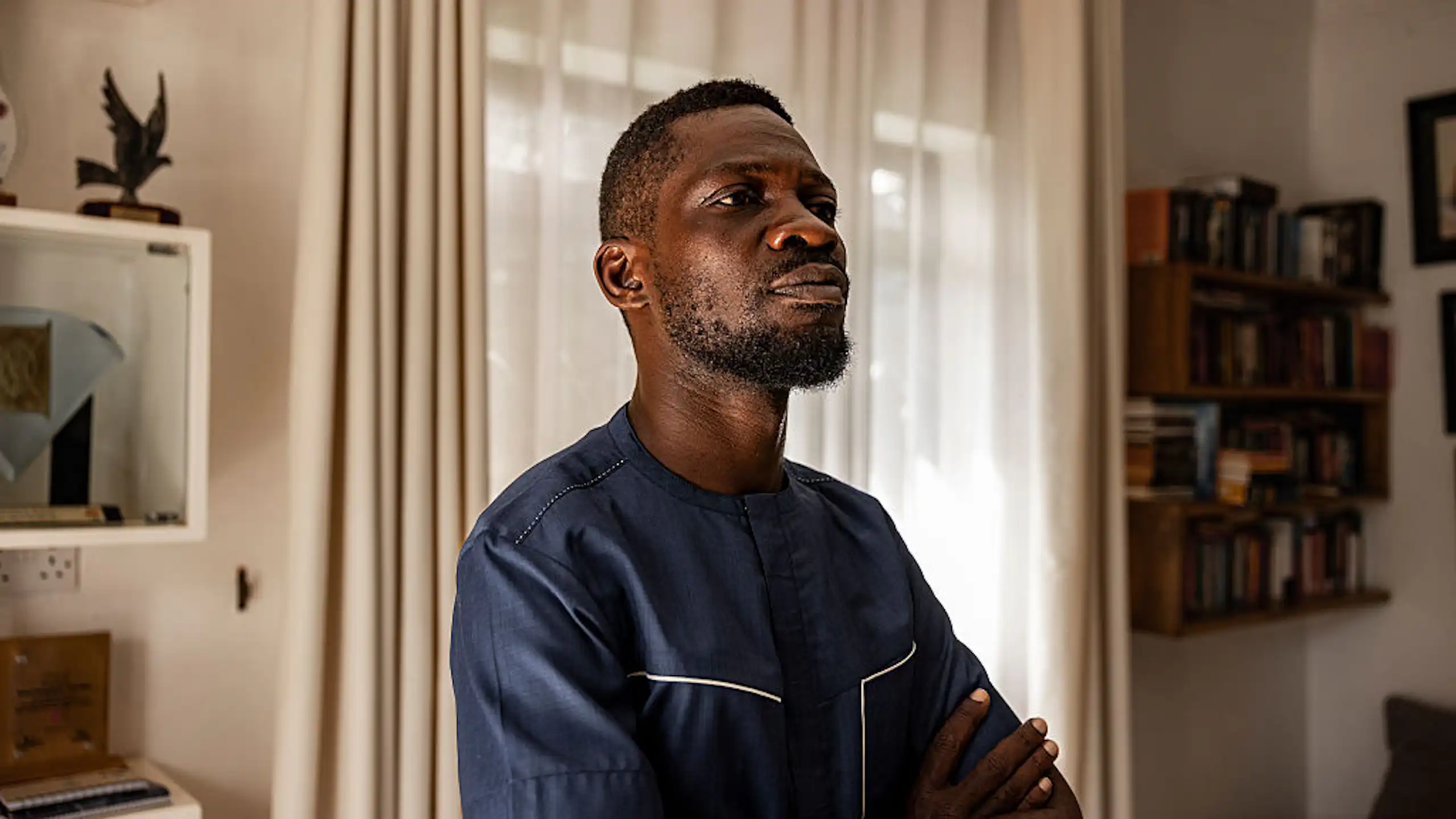 A young African man with a beard stands, armed crossed, in a room with a shelf of books in the background.