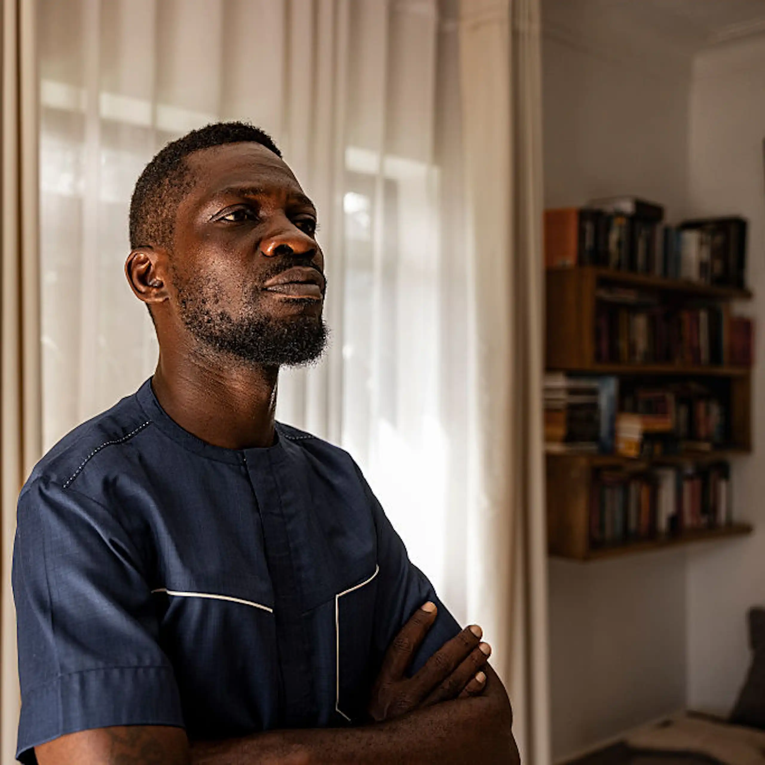 A young African man with a beard stands, armed crossed, in a room with a shelf of books in the background.